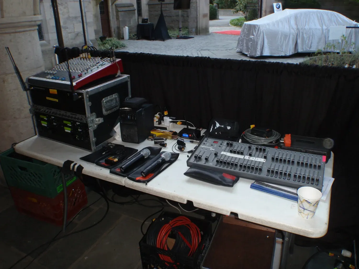 Audio equipment setup with a sound mixer, wireless microphone receivers, two handheld microphones and a lighting console on a table at an outdoor event.