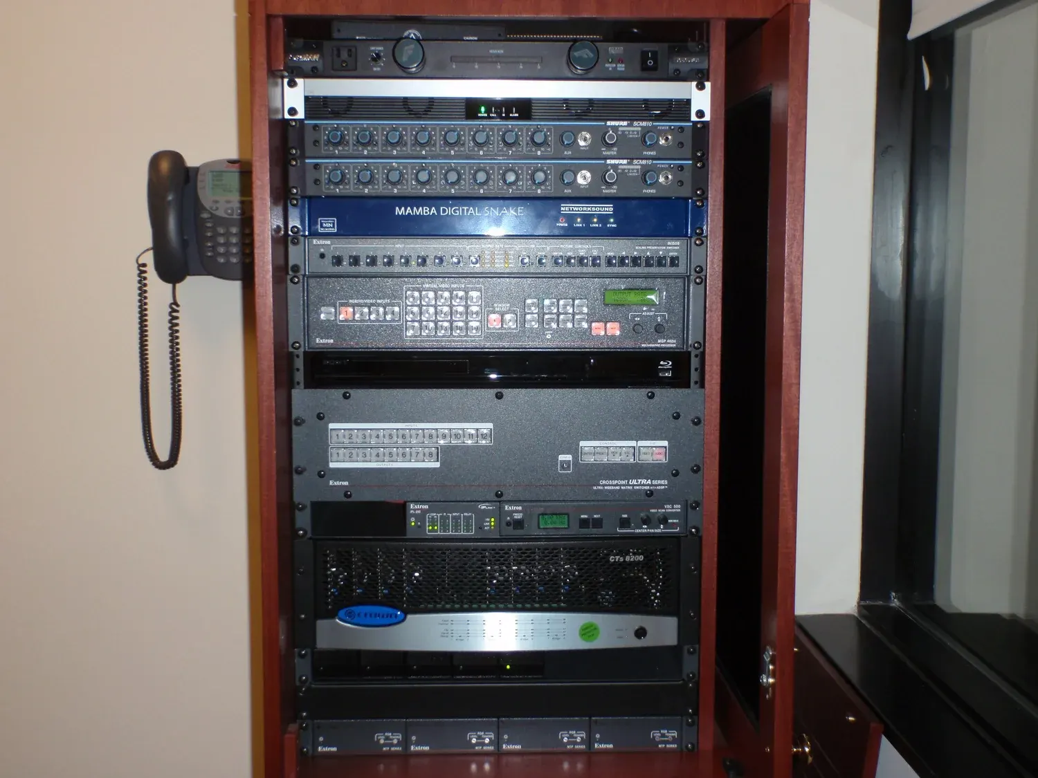 An installed equipment rack in a wood cabinet holding audio mixers, a video switcher, a matrix switcher and power amplifiers, with a wall phone beside it.