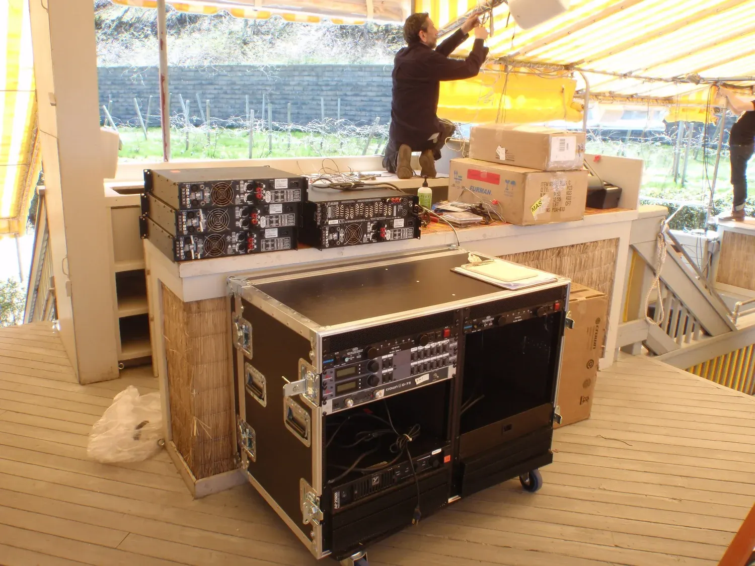 Open road cases of audio equipment being unpacked under a tent, with a technician working at a table behind them.